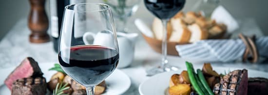 2 Gläser Cabernet Sauvignon auf gedecktem Tick mit Steaks und Beilagen.