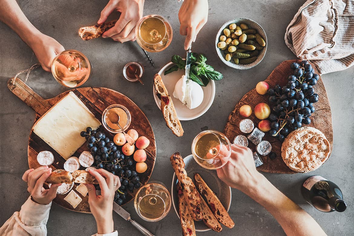Foto von einer Fläche, auf der Wein und Käseplatten stehen, einiege Snacks wie Oliven und Leute, die sich an allem bedienen