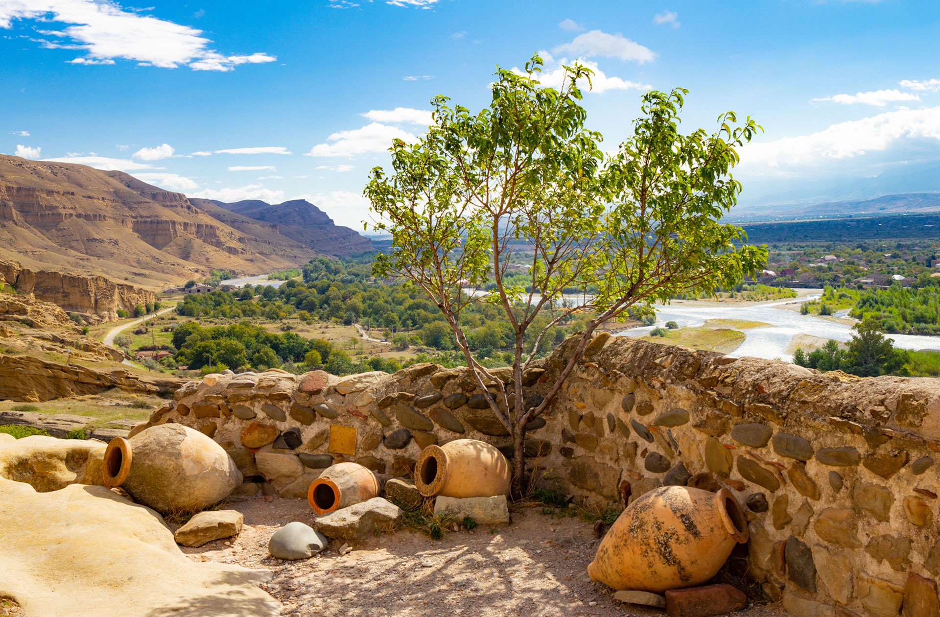 weine aus georgien qvevris draussen blauer himmel Schöner Blick auf den Nationalkrug und einen Olivenbusch vor dem Hintergrund des Tals des Flusses Kura in der antiken Höhlenstadt Uplistsikhe, in der Nähe von Gori, Shida Kartli, Georgien, an einem sonnigen Sommertag.