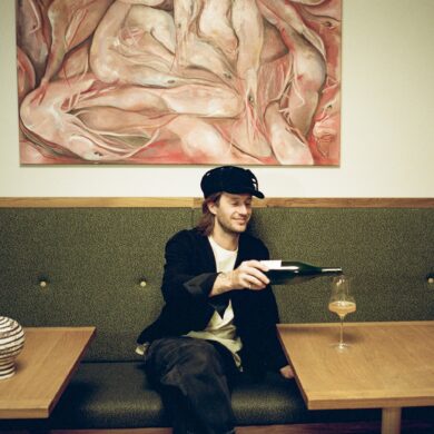 Junger Mann in Weinbar schenkt Naturwein in ein Glas ein, sitzt auf gepolsterter Bank unter expressivem Gemälde; moderne, gemütliche Atmosphäre in einer Weinbar in Wien.