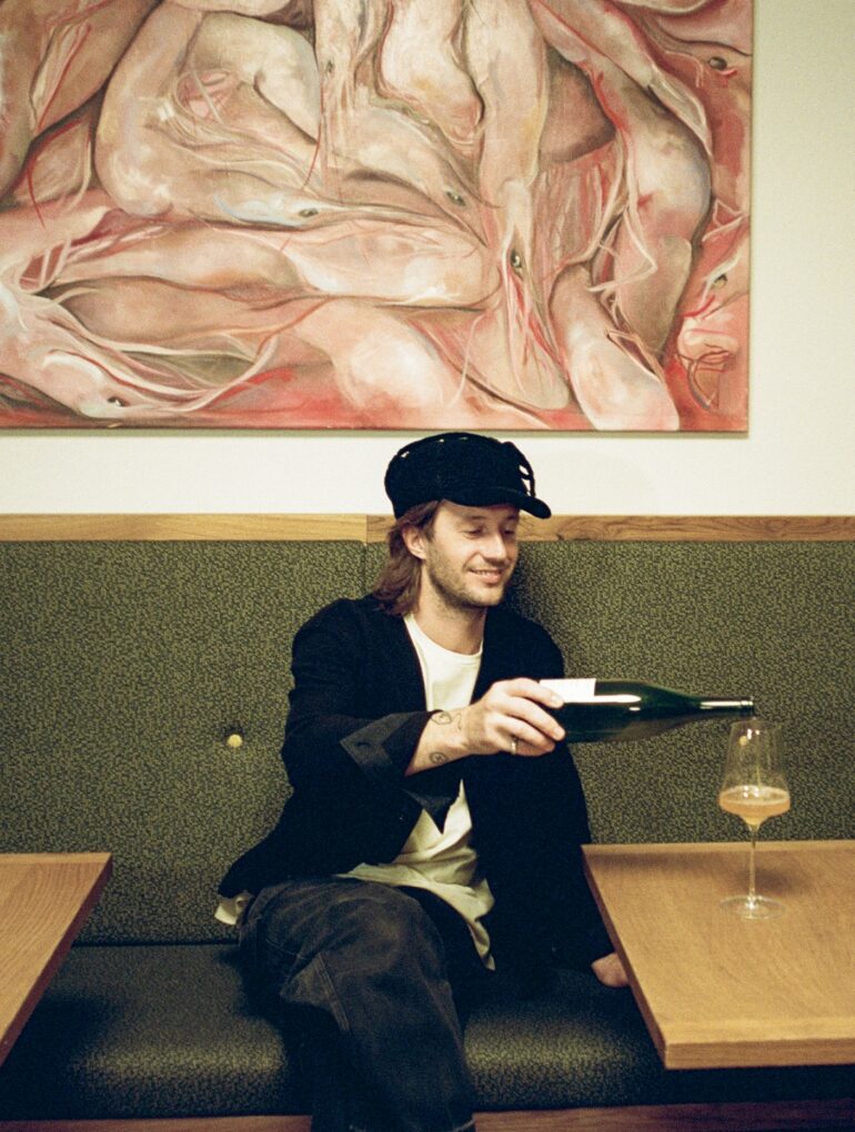 Junger Mann in Weinbar schenkt Naturwein in ein Glas ein, sitzt auf gepolsterter Bank unter expressivem Gemälde; moderne, gemütliche Atmosphäre in einer Weinbar in Wien.