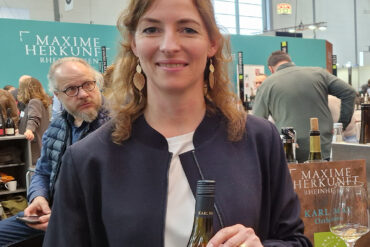 karl may gespraech mit jasmin may prowein 2026 weinblatt 1080x1080 1 Weingut Karl May: Jasmin May mit einer Flasche Weißwein an einem Weingut-Stand auf der Prowein 2026