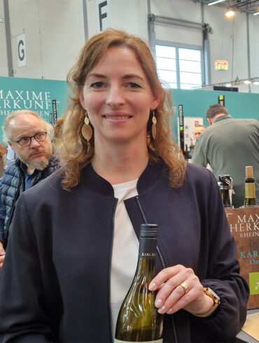 karl may gespraech mit jasmin may prowein 2026 weinblatt 1080x1080 1 Weingut Karl May: Jasmin May mit einer Flasche Weißwein an einem Weingut-Stand auf der Prowein 2026