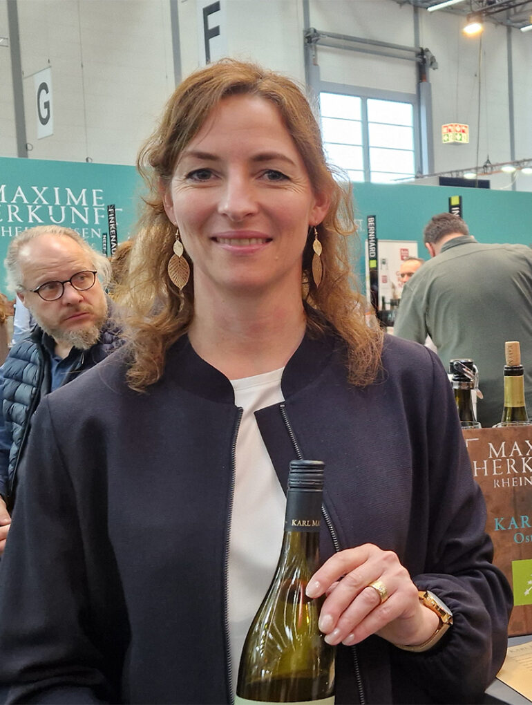 karl may gespraech mit jasmin may prowein 2026 weinblatt 1080x1080 1 Weingut Karl May: Jasmin May mit einer Flasche Weißwein an einem Weingut-Stand auf der Prowein 2026