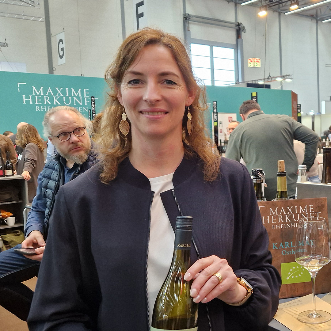 karl may gespraech mit jasmin may prowein 2026 weinblatt 1080x1080 1 Weingut Karl May: Jasmin May mit einer Flasche Weißwein an einem Weingut-Stand auf der Prowein 2026