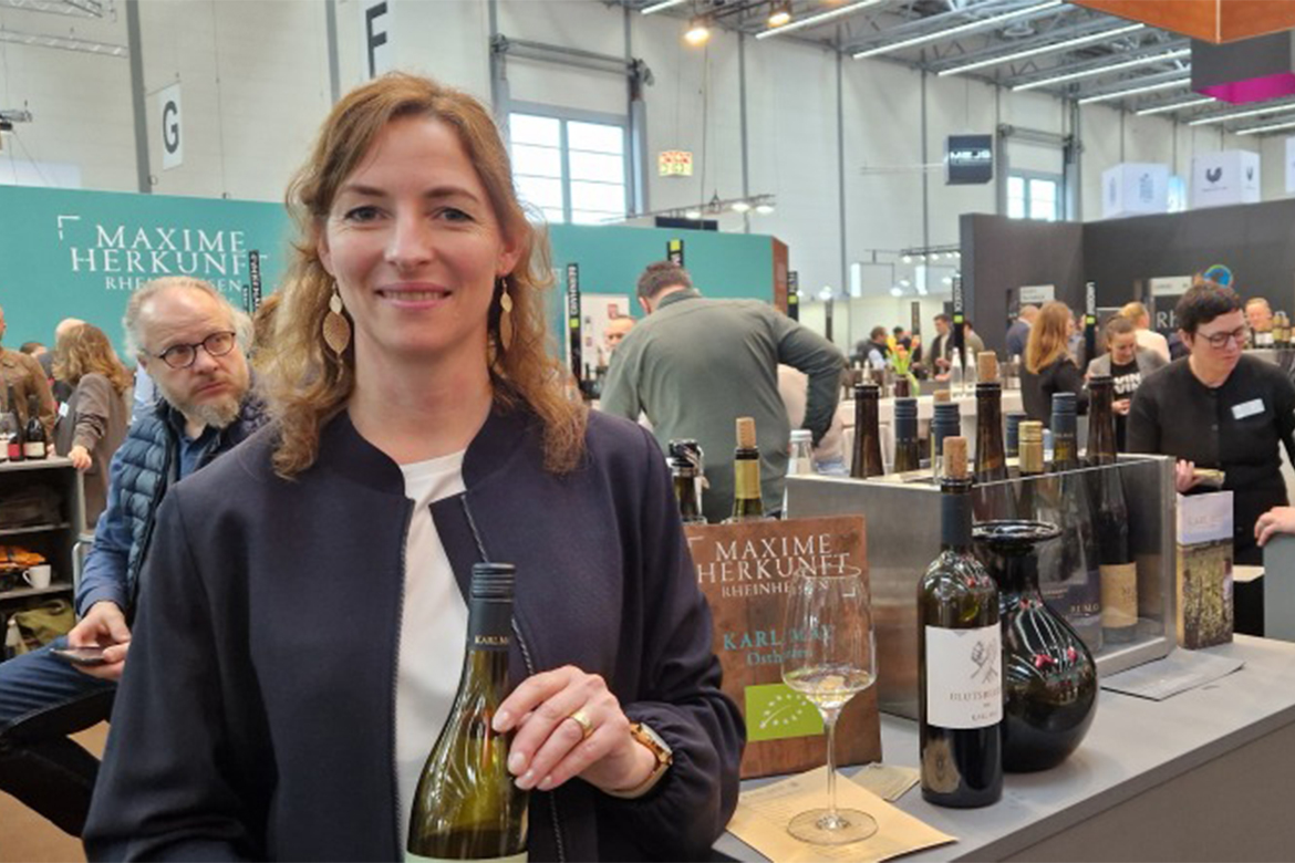 karl may weinblatt bild 3 1170x780 1 Weingut Karl May: Jasmin May mit einer Flasche Weißwein an einem Weingut-Stand auf der Prowein 2026