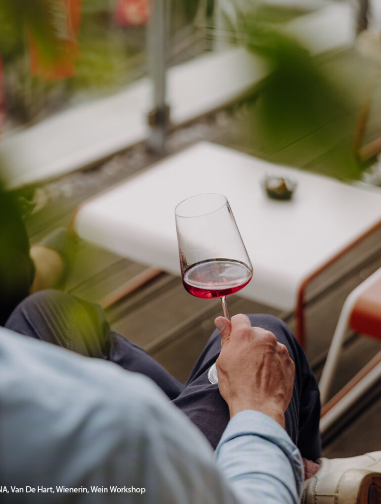 swk blogbeitrag oesterreich wein 1080x1080 1 Österreich Weine entdecken: Person sitzt auf einer Terrasse und hält ein Glas Roséwein, im Hintergrund unscharfe Pflanzen, ein weißer Tisch und rote Fahnen. | KI-generiert